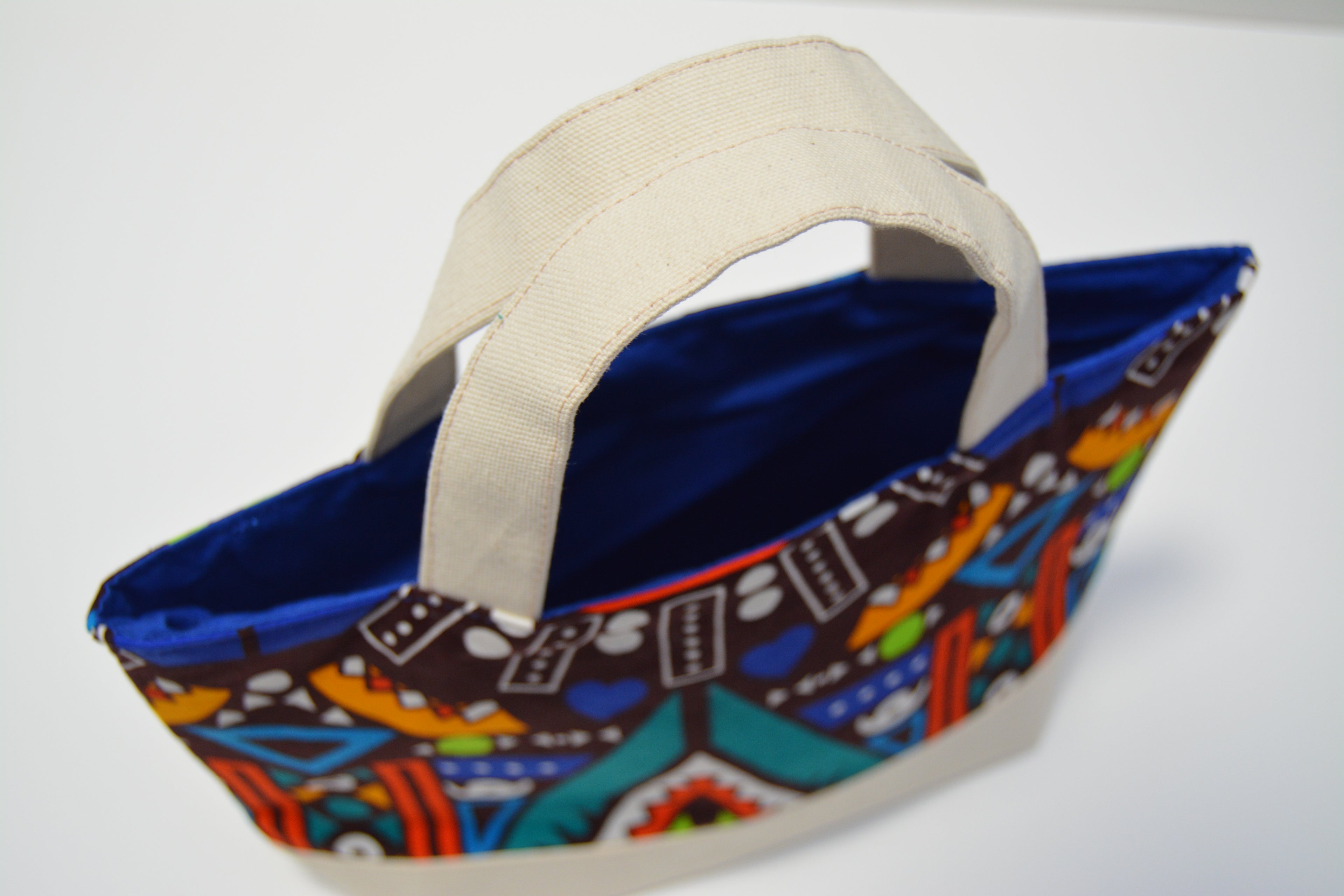 A medium tote bag with a colorful geometric patterned exterior, a blue fabric lining, and thick cream canvas handles, shown on a plain white background.