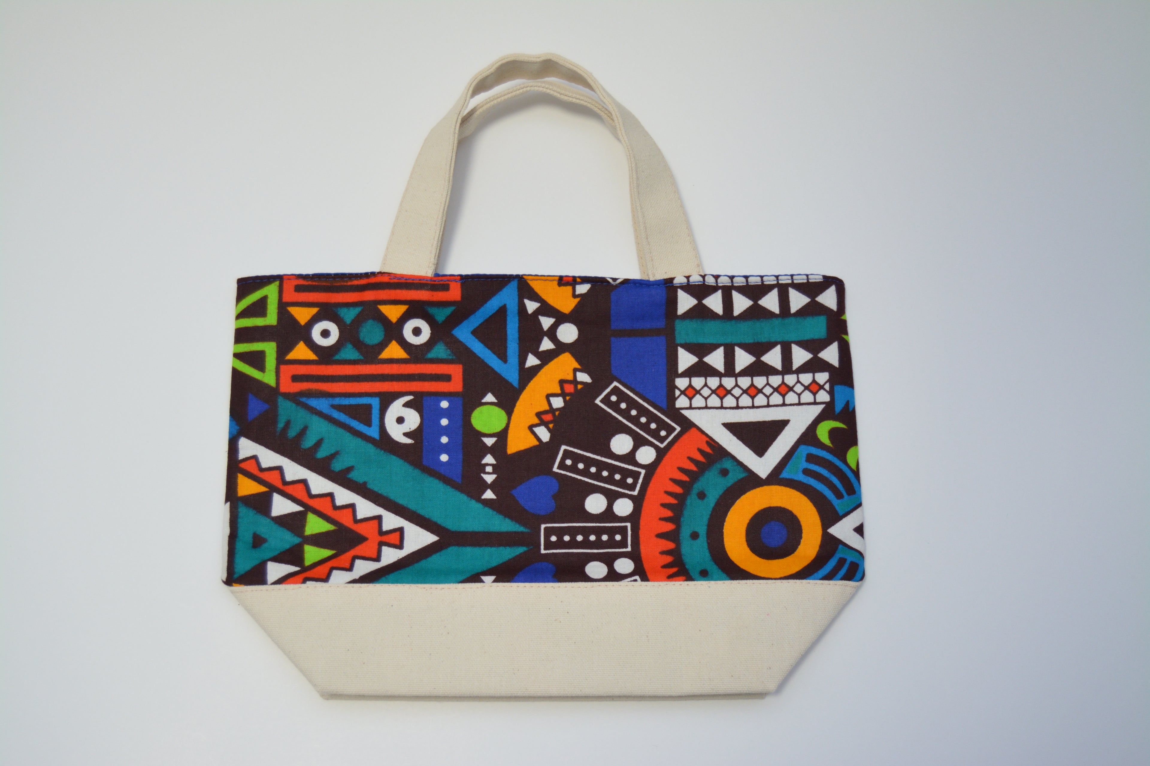 A canvas tote bag with a colorful geometric pattern on the front panel and solid cream-colored handles and base, photographed against a light gray background.