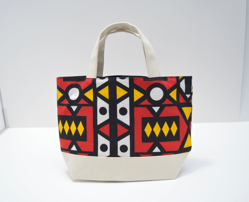 A canvas tote bag with a cream-colored base and handles, featuring a bold geometric tribal pattern in red, black, yellow, and white on the upper body, set against a plain white background.