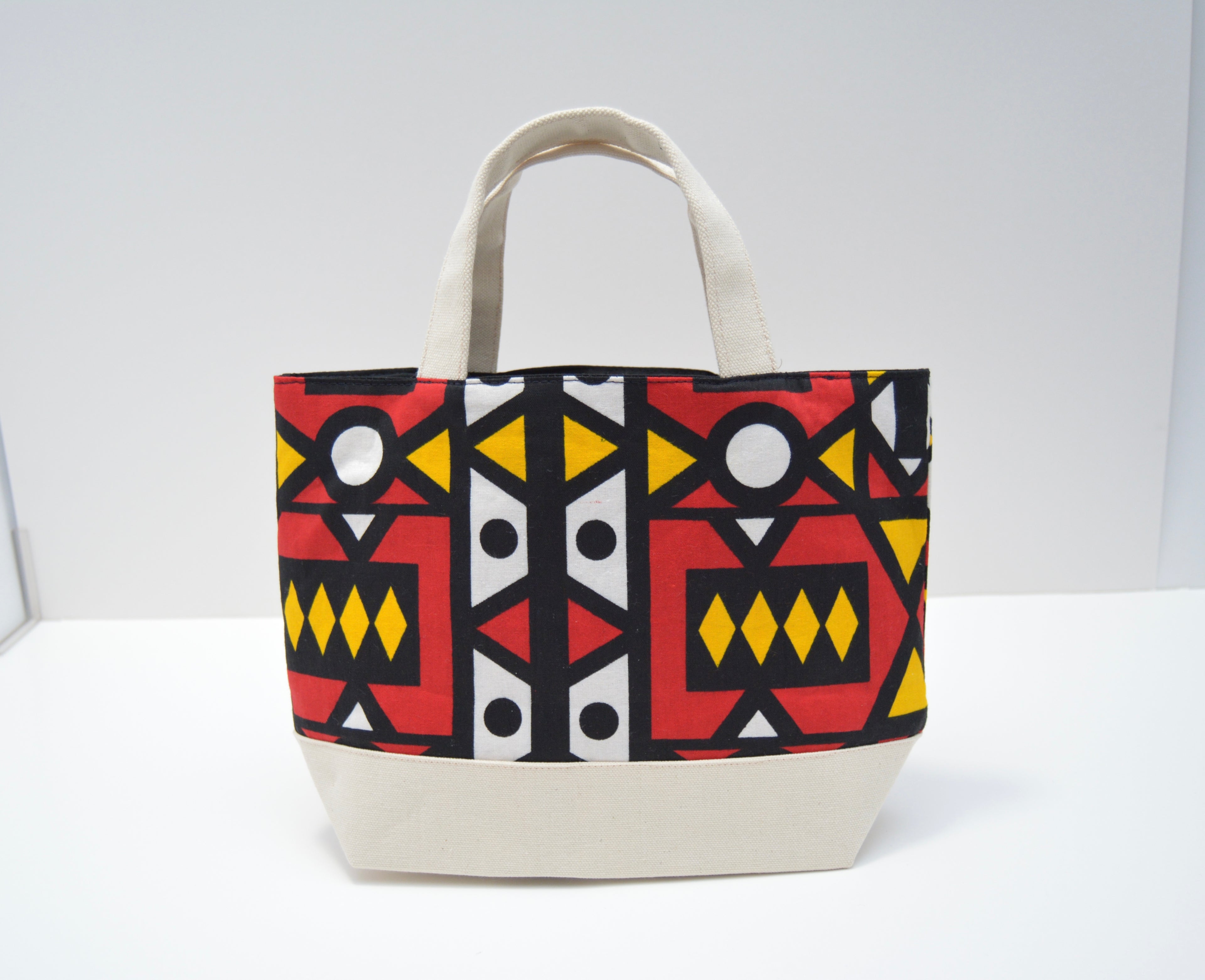 A canvas tote bag with a cream-colored base and handles, featuring a bold geometric tribal pattern in red, black, yellow, and white on the upper body, set against a plain white background.