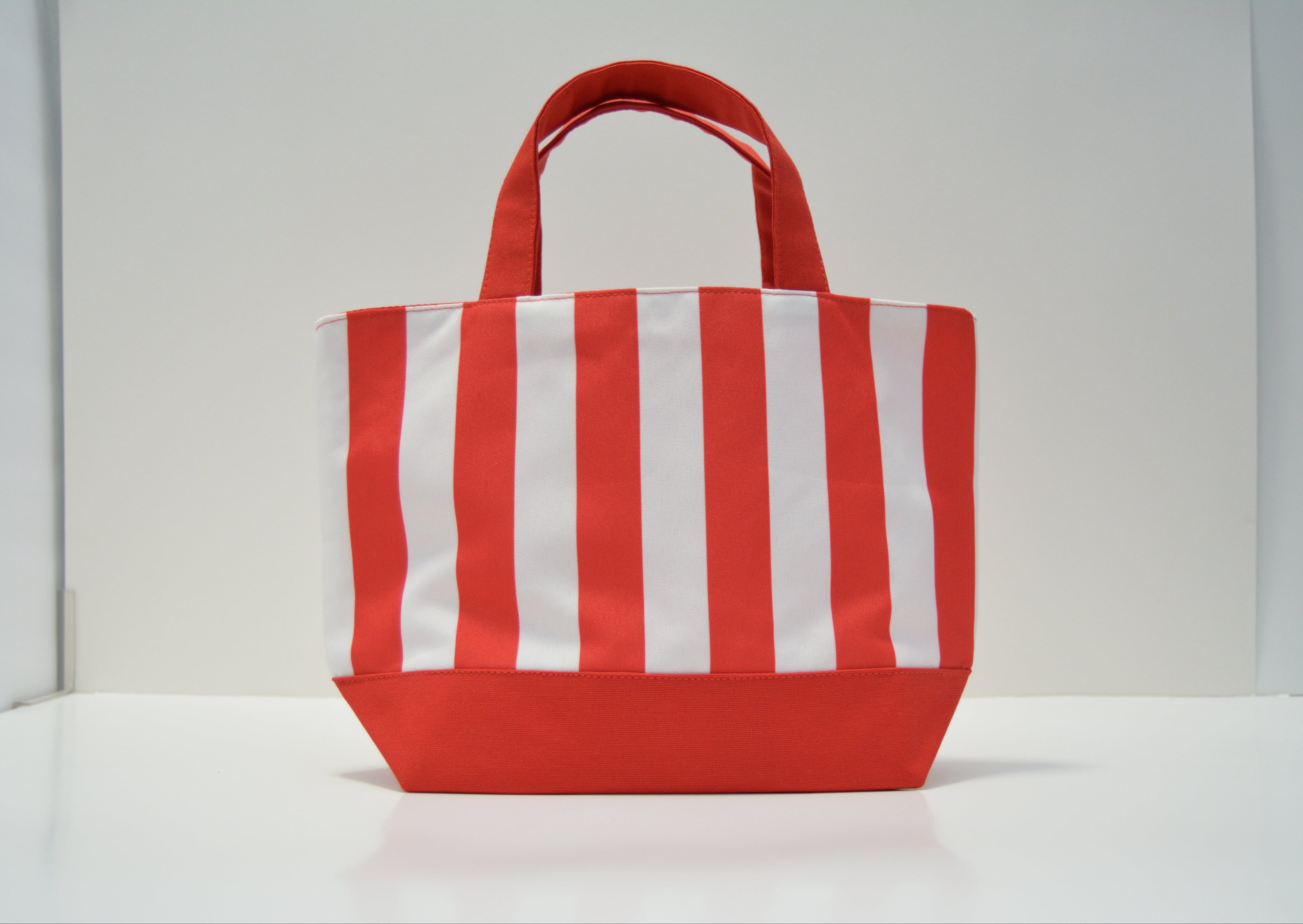 A red and white vertically striped canvas tote bag with a solid red base and top handles, displayed on a clean white surface against a white background.