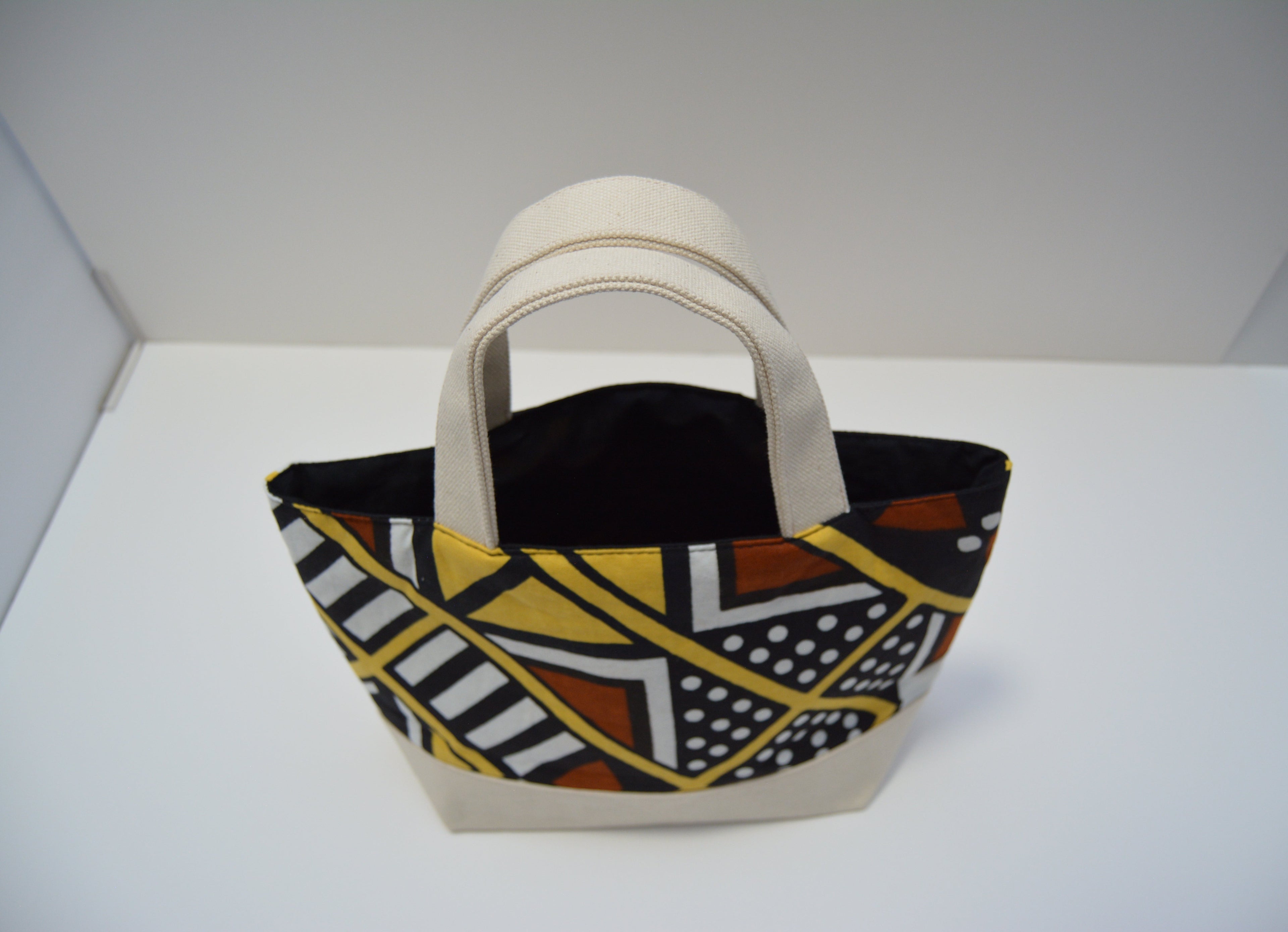 A small tote bag with cream-colored handles and base, featuring a bold African wax print pattern in yellow, black, white, and brown on the front panels, set against a plain white background.