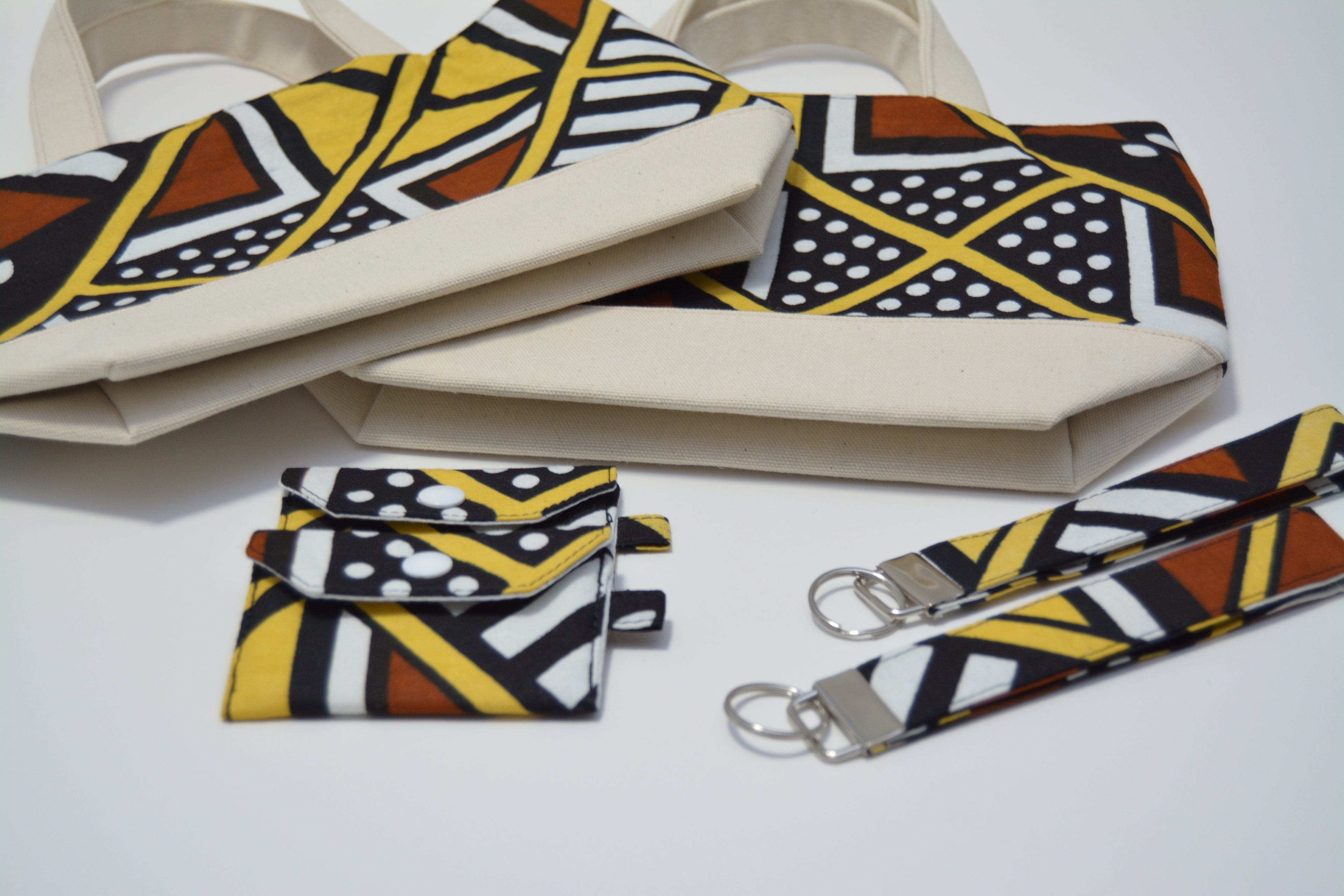 A matching fabric accessory set featuring a canvas tote bag, a small pouch, and two wristlets, all decorated with a bold African wax print pattern in yellow, brown, black, and white, with silver metal keyrings on two of the items.