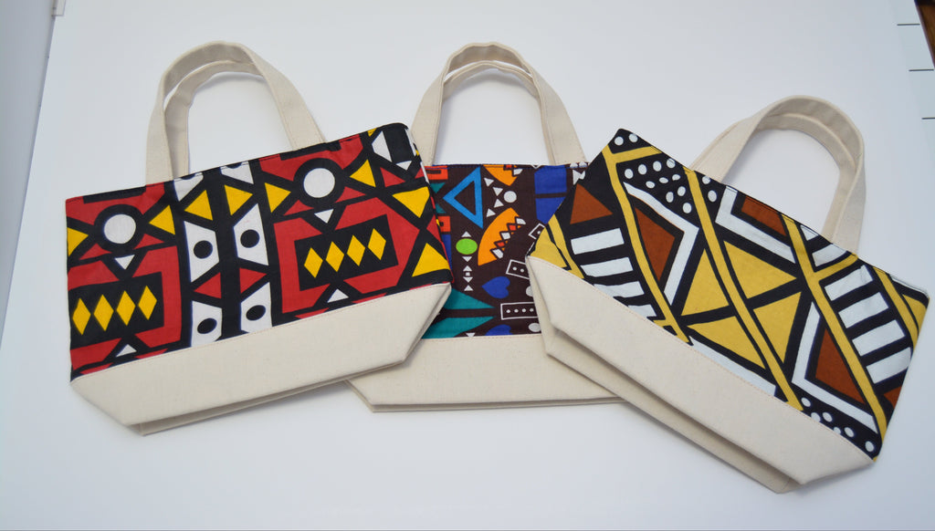 Three canvas tote bags with cream-colored handles and bases, decorated with vibrant African wax print patterns in red, yellow, black, white, and blue, arranged on a white surface.